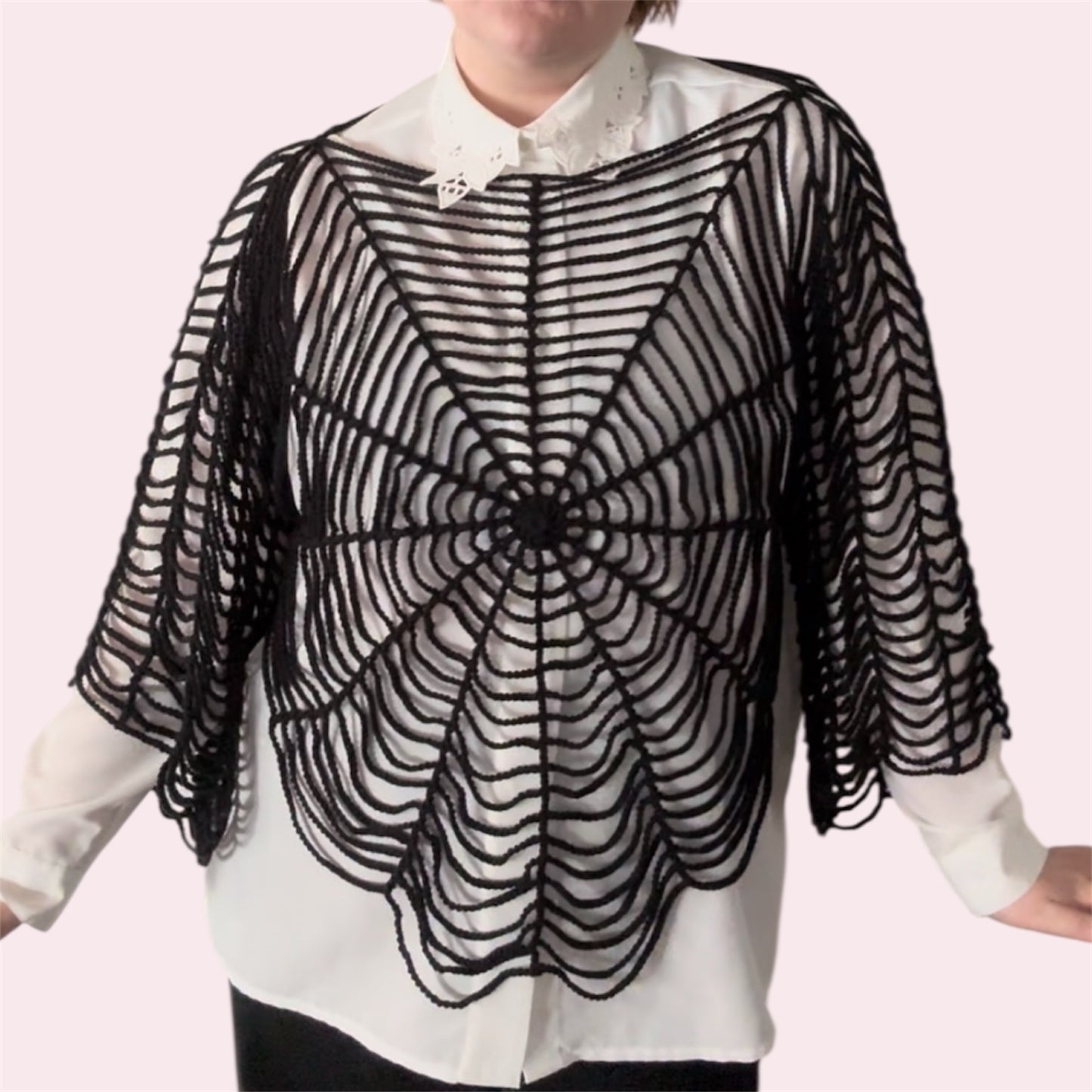 Person wearing a black spider web patterned garment over a white shirt on a pink background